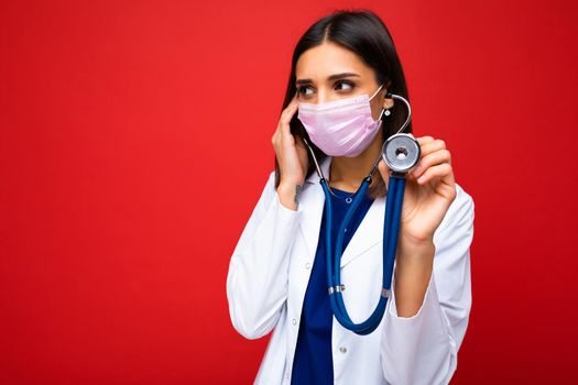 Photo of professional confident young european brunnet woman doctor in medical mask and white coat, stethoscope over neck, ready help patient, fight disease. Free space