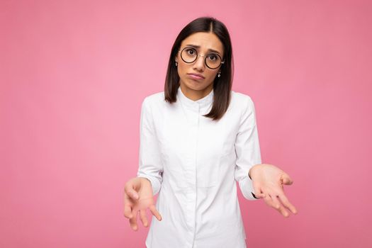 Photo shot of beautiful sorrowful young brunette woman wearing white shirt and stylish optical glasses isolated over pink background looking at camera.