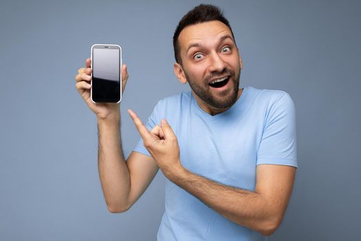 Amazed handsome young unshaven brunet man wearing everyday blue t-shirt isolated over blue background holding and showing mobile phone with empty display for cutout looking at camera and pointing finger at smartphone.