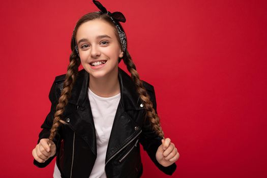 Charming positive smiling brunette little female teenager with pigtails wearing stylish black leather jacket and white t-shirt standing isolated over red background wall looking at camera.