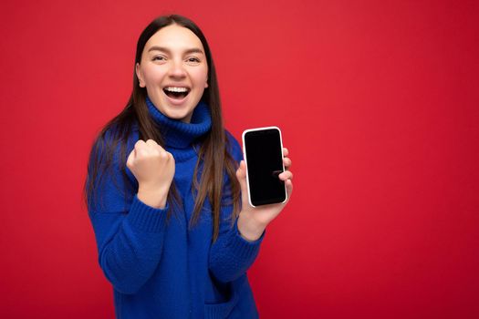 beautiful happy young brunette woman wearing casual blue sweater isolated over red background with empty space holding in hand mobile phone and showing smartphone with empty screen for mockup looking at camera.