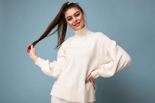 Young beautiful happy positive smiling stylish brunette woman wearing casual white sweater poising isolated on blue background wall.