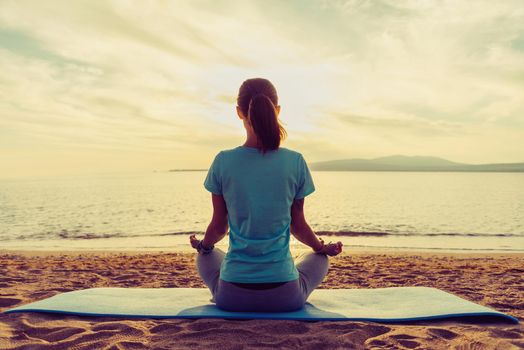 Young woman meditating in pose of lotus on beach near the sea at sunset in summer, rear view