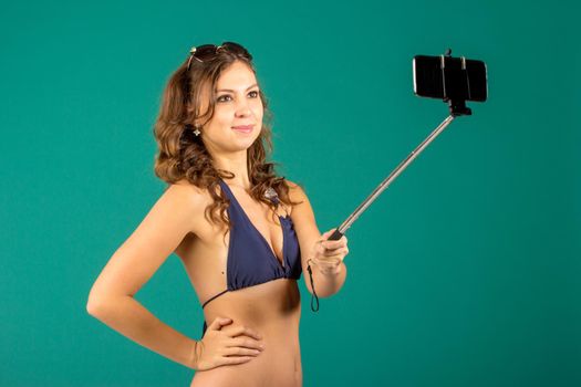 Happy young woman in blue bikini swimsuit taking selfie with smatphone on green background