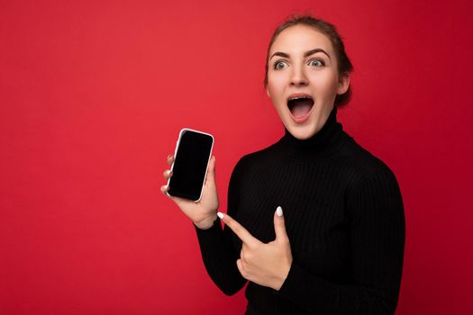 Photo of Shocked attractive positive young brunette woman wearing black sweater standing isolated over red background showing mobile phone with empty screen for mockup looking to the side pointing finger at gadjet.