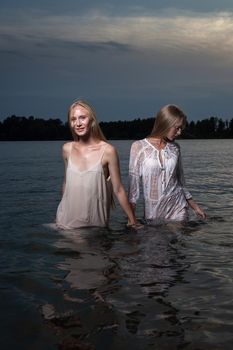 two attractive young twin sisters with long blond hair posing in light dresses in water of lake at summer night. stylish fashion photoshoot with flashlight. pretty models outdoors evening photosession