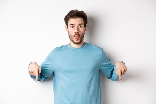 Amazed young man with beard, standing in casual blue shirt, saying wow and pointing down, look here gesture, standing over white background.