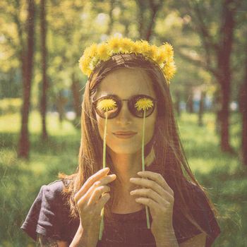 Smiling beautiful girl in wreath of yellow dandelions closed her eyes with dandelions in summer park, concept of happiness and summer mood. Image with old textured effect