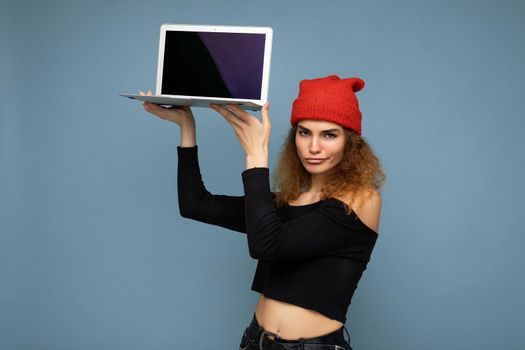 Close-up portrait of beautiful funny self-confident dark blond curly young lady with dack lips holding laptop computer looking at camera wearing black crop top and red and orange do-rag isolated over light blue wall background. Mock up, copy space, empty space