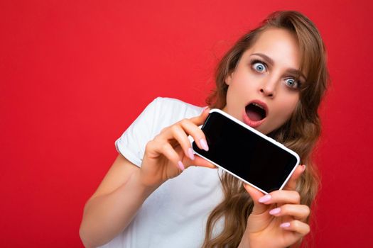 Surprised shocked beautiful smiling young blonde woman good looking wearing white t-shirt standing isolated on red background with copy space holding phone showing smartphone in hand with empty screen display for mockup looking at camera.