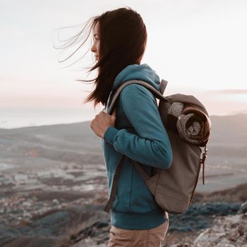 Hiker young woman with backpack standing on peak of mountain and looking into the distance in summer at sunset
