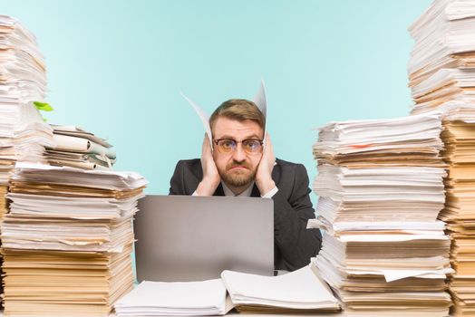 Shocked businessman sitting at the table with many papers in office, he is overloaded with work - image