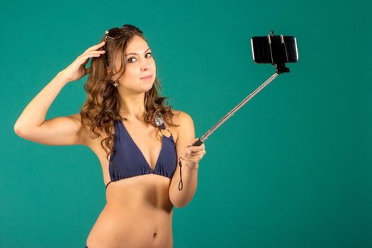 Happy young woman in blue bikini swimsuit taking selfie with smatphone on green background