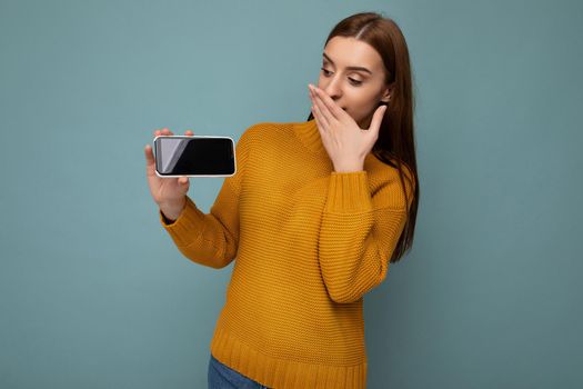 Amazed attractive positive young woman wearing orange sweater poising isolated on blue background with empty space holding in hand and showing mobile phone with empty space for mockup looking at gadjet display.