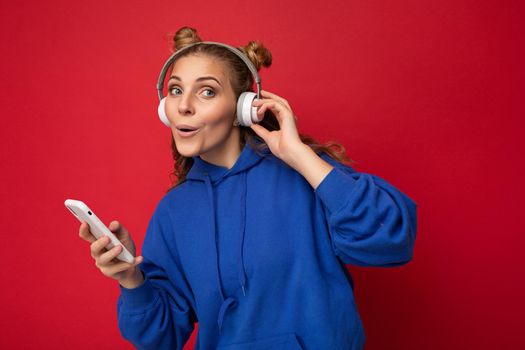 Photo of beautiful joyful funny young woman wearing stylish blue hoodie isolated over red background wearing white bluetooth wireless headphones and listening to music and using mobile phone surfing on the internet looking at camera.