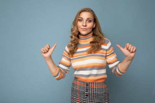 Photo portrait of young pretty beautiful happy self-confident blonde woman with sincere emotions wearing striped pullover isolated on blue background with empty space and pointing fingers at herself.