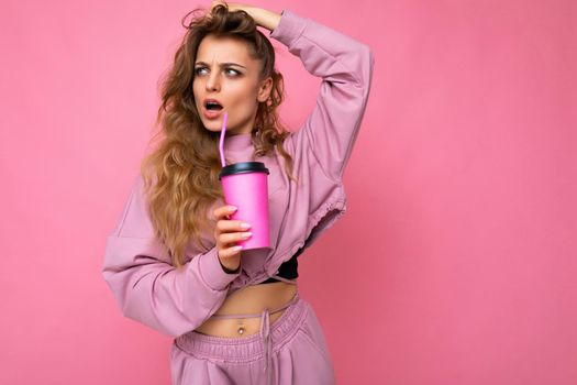 Photo shot of beautiful young shocked blonde woman wearing pink sport clothes isolated over pink background holding paper coffee cup for mockup drinking and looking to the side.