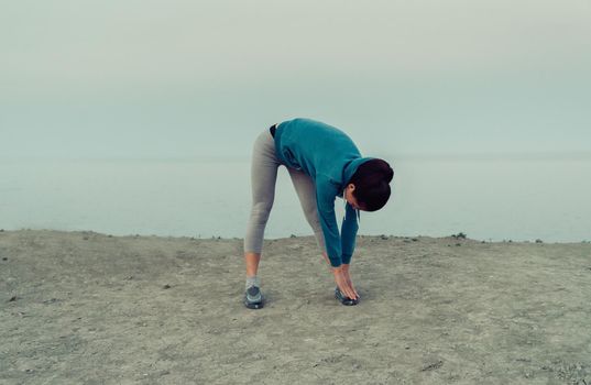 Young sporty woman stretching and preparing to run on coast in summer in the morning. Concept of sport and healthy lifestyle