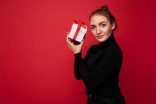 Charming positive young brunette woman isolated over red background wall wearing black sweater holding white gift box with red ribbon and looking at camera. Empty space