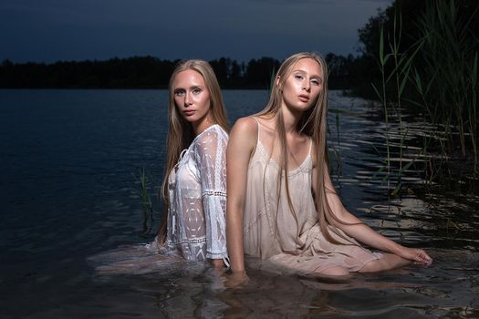 two young twin sisters with long blond hair posing in light dresses in water of lake at summer night, looking at camera. stylish fashion photoshoot with flashlight. outdoors evening photosession