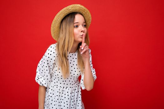 Photo shot of young beautiful blonde woman wearing casual dress and straw hat isolated over red background with copy space showing shh geasture. Be quiet concept.