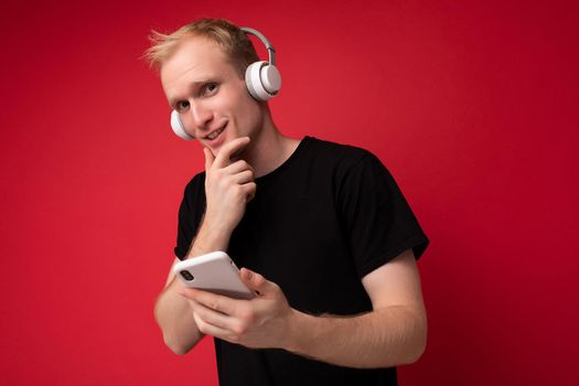 Thinking handsome blonde young man wearing black t-shirt and white earphones standing isolated over red background with copy space holding smartphone and communicating online listening to music and having fun looking at camera.