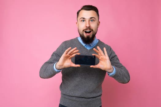 Handsome happy cool young brunette unshaven man with beardwearing stylish grey sweater and blue shirt standing isolated over pink background wall holding smartphone and showing phone with empty screen display looking at camera.