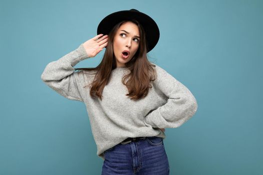 Portrait of young emotional beautiful brunette woman with sincere emotions wearing stylish grey pullover and black hat isolated over blue background with copy space and listening to gossip with shock expression.