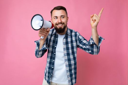 Shot of young handsome positive happy smiling brunet man with beard with sincere emotions wearing casual t-shirt and stylish check shirt isolated on pink background with copy space and speaking into magaphone to announce something, showing one finger to pay attention.