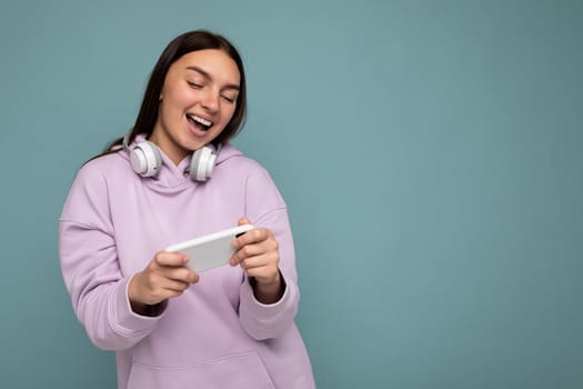 Photo shot of beautiful joyful smiling young female person wearing stylish casual outfit isolated over colorful background wall wearing white bluetooth wireless earphones and listening to music and using mobile phone looking at gadjet display. copy space