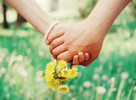 Young loving couple holding hands each other with bouquet of yellow dandelions in summer park, view of hands