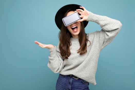 Pretty young smiling woman wearing black hat and grey sweater holding phone looking at camera isolated on background. mock up