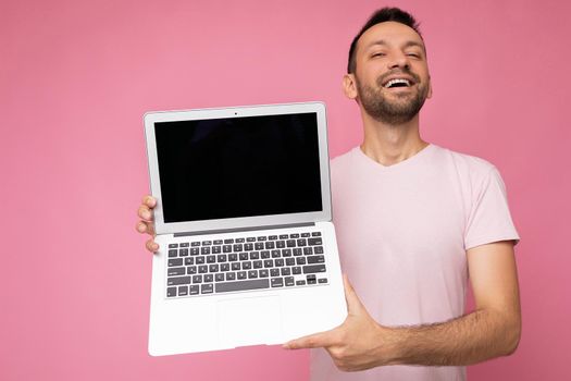 Handsome smiling brunet man holding laptop computer looking at camera in t-shirt on isolated pink background.