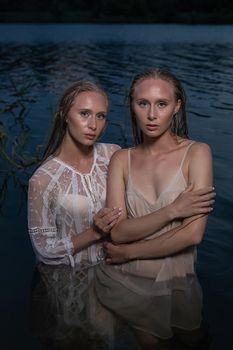 two young twin sisters with long blond hair posing in light dresses in water of lake at summer night, looking at camera. stylish fashion photoshoot with flashlight. outdoors evening photosession