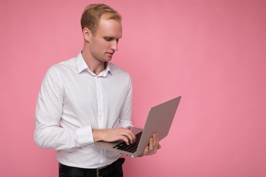 Side profile photo shot of handsome confident blonde man holding computer laptop typing on keyboard wearing white shirt looking at monitor isolated over pink background.