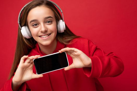 Closeup portrait of amazibg beautiful brunette girl wearing red hoodie isolated on red background holding and showing smartphone with empty display for cutout wearing white wireless headsets listening to energetic music looking at camera.