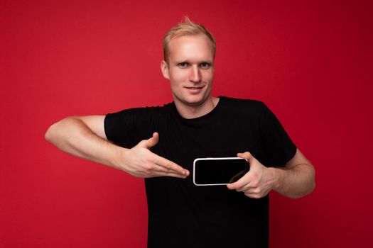 Photo of handsome smiling adult male person good looking wearing casual outfit standing isolated on background with copy space holding smartphone showing phone in hand with empty screen display for mockup pointing at gadjet looking at camera.