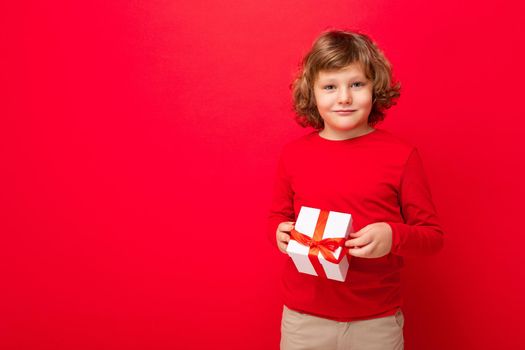 Shot of happy positive blond curly boy standing isolated over red background wall wearing red sweater holding gift box and looking at camera. Empty space