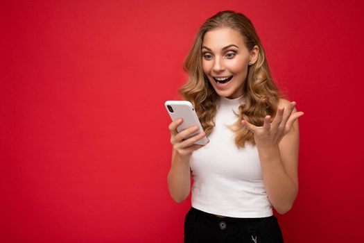 Photo of attractive crazy amazed surprised young woman wearing casual stylish clothes standing isolated over background with copy space holding and using mobile phone looking at device display.