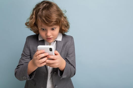 Photo of Handsome shocked boy with curly hair wearing grey suit holding and using phone isolated over blue background looking at smartphone communicating vie sms.