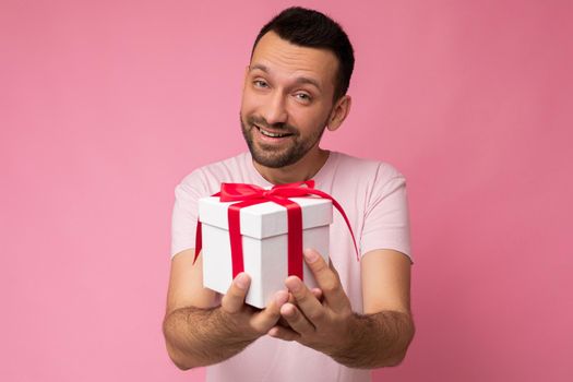 Photo shot of handsome positive brunette unshaven young man isolated over pink background wall wearing pink t-shirt holding white gift box with red ribbon and looking at camera.