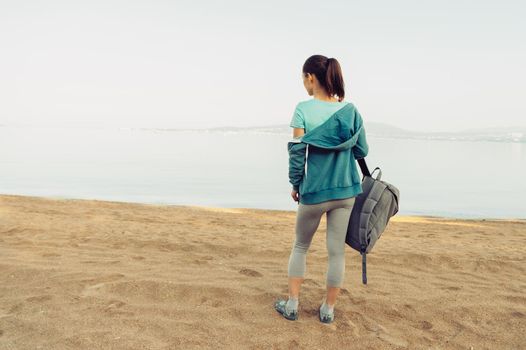 Sporty young woman with backpack standing on beach in summer and ready for workout. Space for text in left part of image