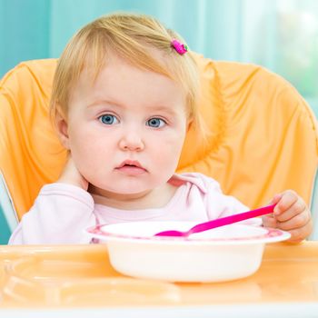 cute one year old girl in a highchair for feeding with a spoon and a plate in the kitchen