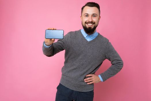 Positive smiling Handsome happy cool young brunette unshaven man with beardwearing stylish grey sweater and blue shirt standing isolated over pink background wall holding smartphone and showing phone with empty screen display looking at camera.