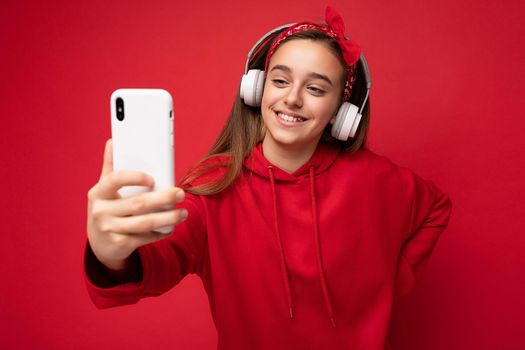 Closeup shot of smiling pretty brunette girl wearing red hoodie isolated on red background holding and using smartphone taking selfie photo wearing white wireless headphones listening to cool music looking at phone display.