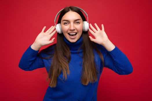 Attractive positive young brunette woman wearing blue sweater isolated over red background wall wearing white bluetooth earphones listening to cool music and having fun looking at camera.