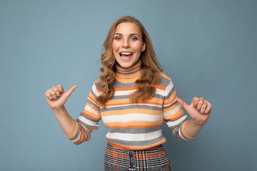 Portrait of young winsome attractive happy smiling blonde woman with wavy-hair wearing striped sweater isolated over blue background with empty space and pointing fingers at herself.