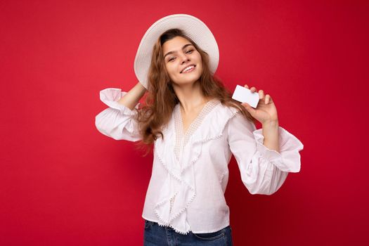 Shot of attractive positive smiling young dark blonde woman wearing white blouse and white hat isolated over red background holding credit card looking at camera.