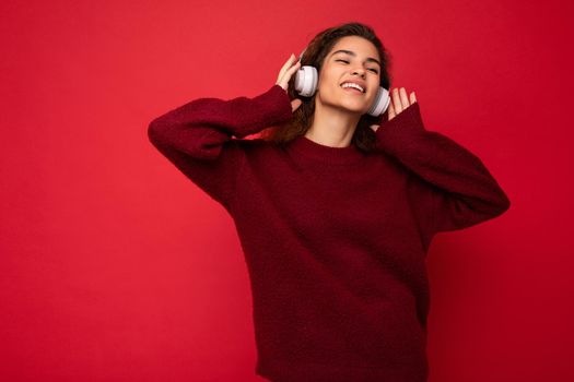 Photo of beautiful happy smiling young brunette curly woman wearing dark red sweater isolated over red background wall wearing white bluetooth headphones listening to music and having fun with close eyes.