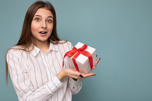 Photo of pretty positive surprised brunette young woman isolated over blue background wall wearing white shirt holding white gift box with red ribbon and looking at camera. Copy space, mockup